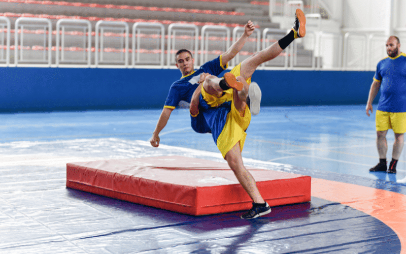 A wrestling coach is instructing a group of wrestlers in a gym, surrounded by mats and wrestling equipment. The athletes are practicing various techniques and drills, while the coach provides guidance and feedback